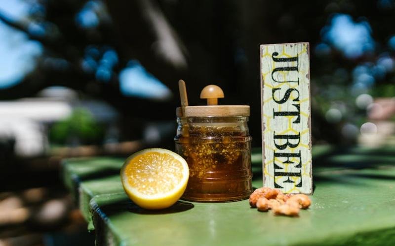 Close-up of a honey jar, lemon, and 'Just Bee' sign on a green table outdoors.