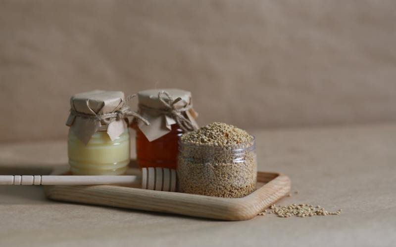 A rustic still life image featuring jars of honey and grains on a wooden tray.