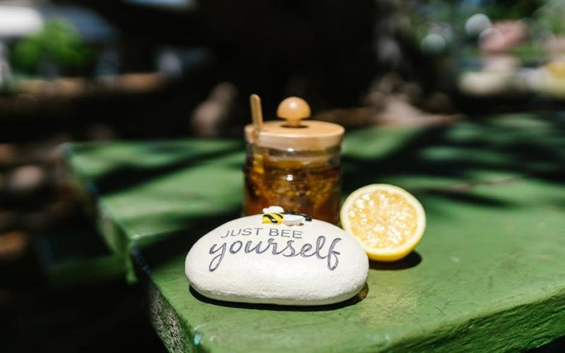 A jar of honey, lemon slice, and stone with 'Just Bee Yourself' outdoors on a sunny day.