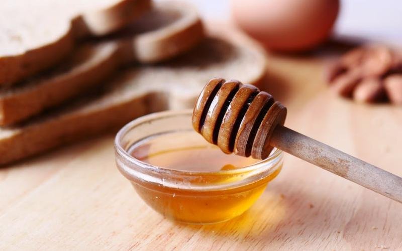 Close-up of honey dipper in a bowl with honey on wooden table beside sliced bread.