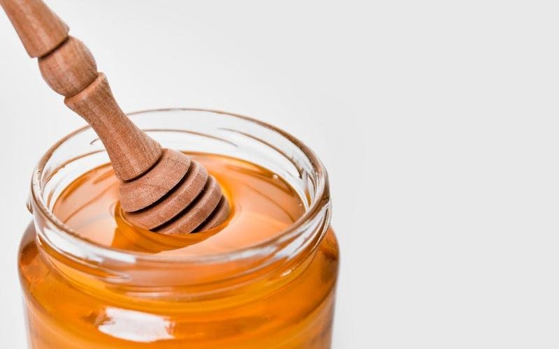 Glass jar of honey with a wooden dipper, set against a white background. Perfect for food and kitchen-related content.