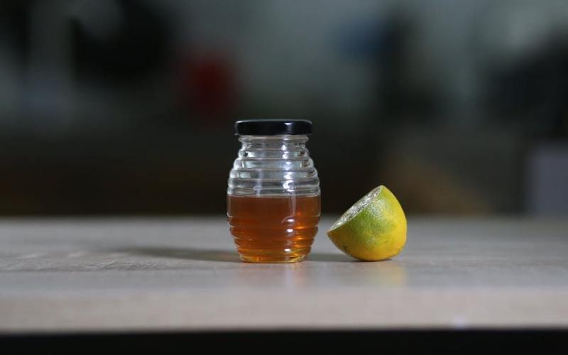 A glass jar filled with honey next to a sliced lemon on a wooden table indoors.