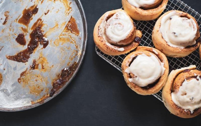 Delicious homemade cinnamon rolls with icing on cooling rack next to an empty baking tray.