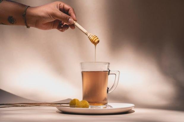 A hand drips honey into a glass mug of tea using a honey dipper, with a warm, inviting atmosphere.