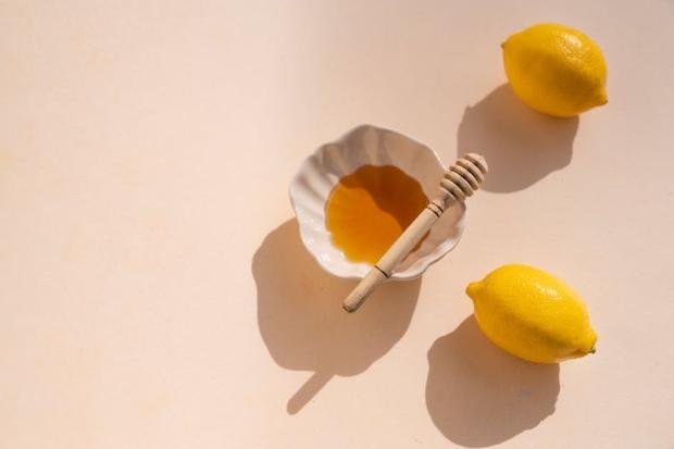 Top view of honey bowl with wooden dipper and lemons on a light background.