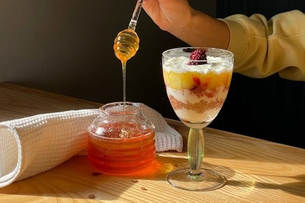 A hand drizzles honey over a fruit parfait in warm sunlight, creating a delicious dessert scene.