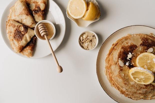 Top view of pancakes with lemon slices, honey, and almonds on a white background.