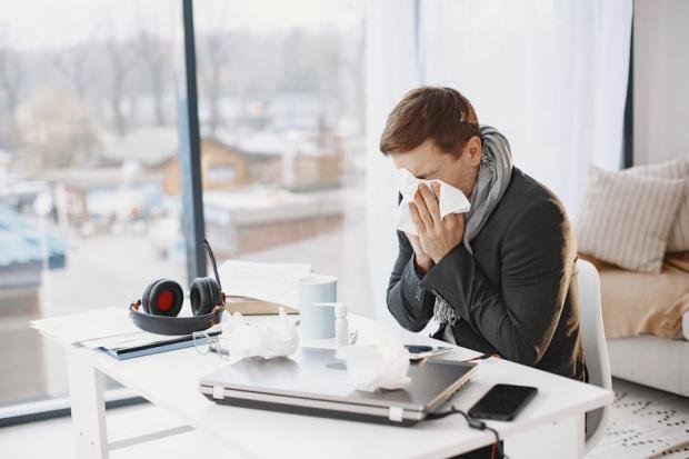 Man sneezing at home desk, indicating remote work during illness.
