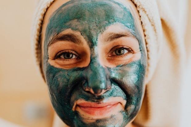 Close-up of a smiling woman with a green face mask, promoting skincare ritual.